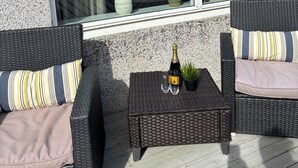 Un balcón con mobiliario de mimbre, una mesa con una botella y vasos, y plantas en macetas.