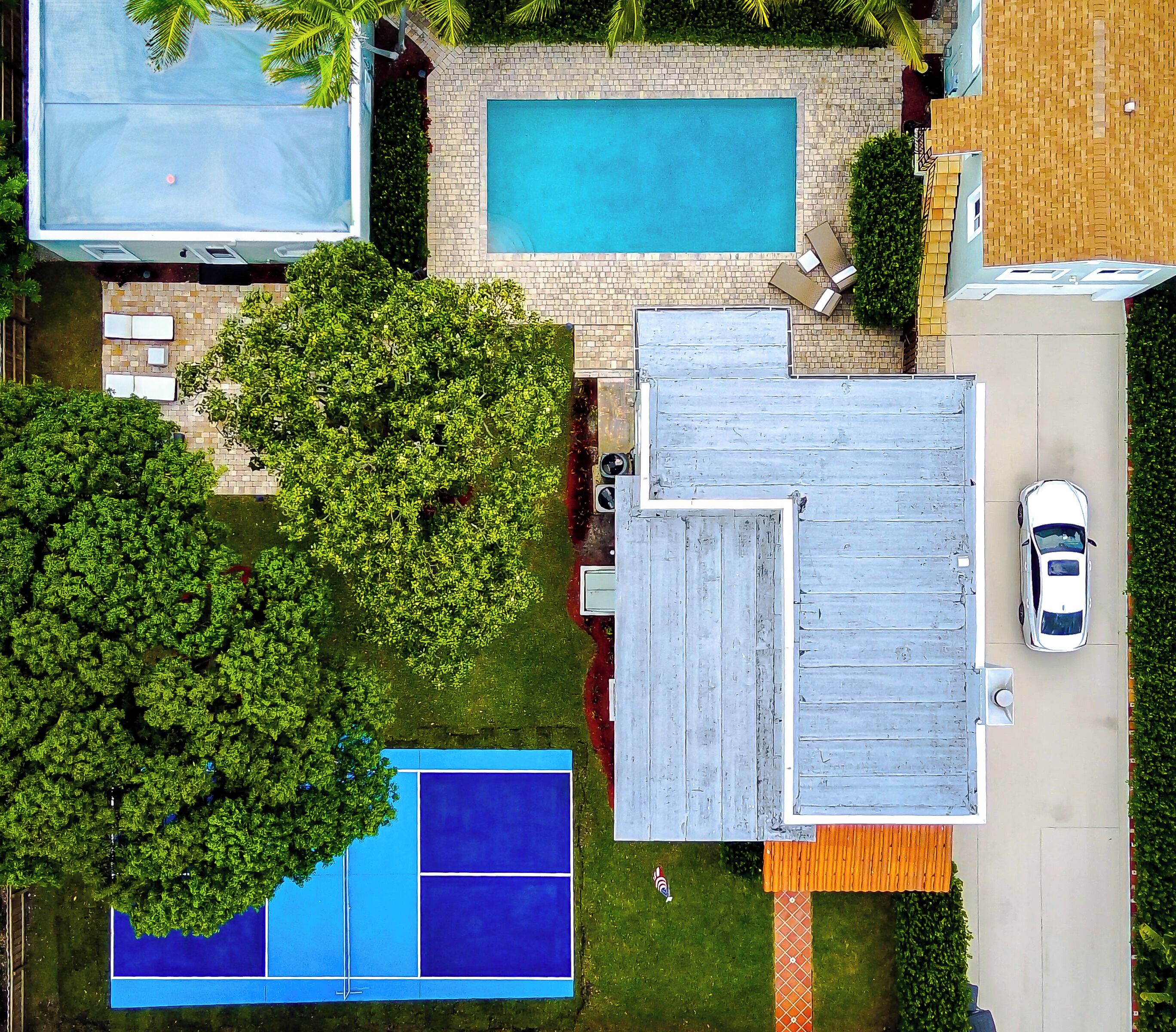 An aerial view of a residential area with a swimming pool, tennis court, and a car parked on the street.