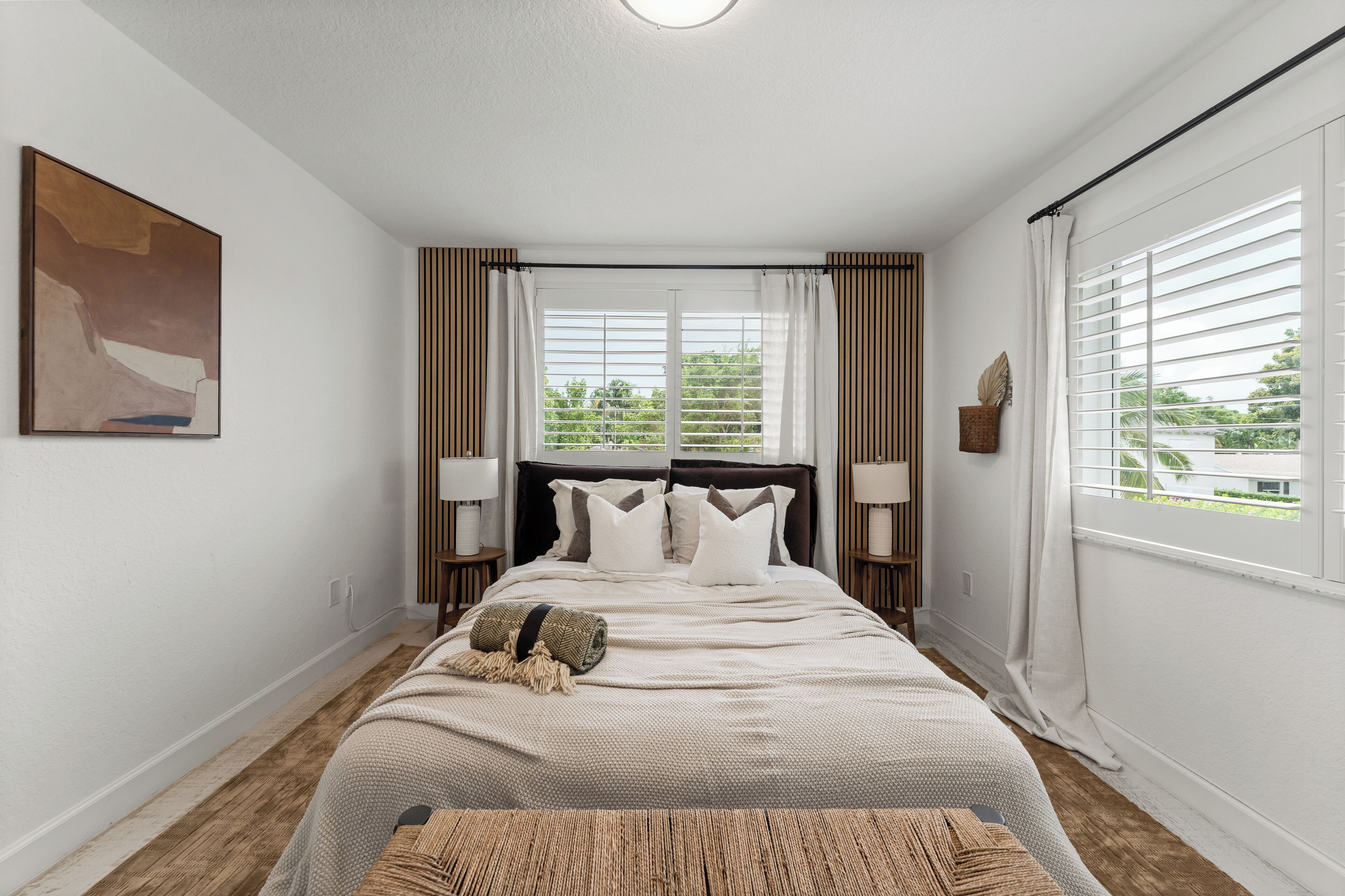 A bedroom with a large bed, two bedside tables with lamps, a window with blinds, and a painting on the wall.