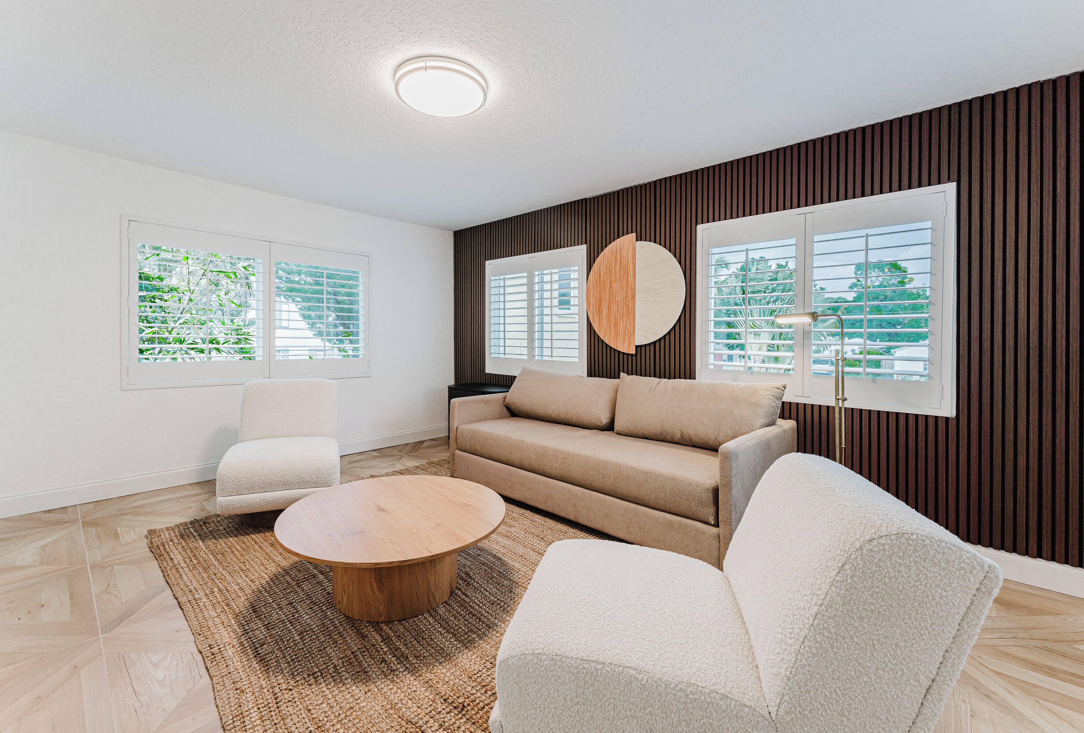 A modern living room with a beige sofa, two white armchairs, a round wooden coffee table, and a large window with blinds.
