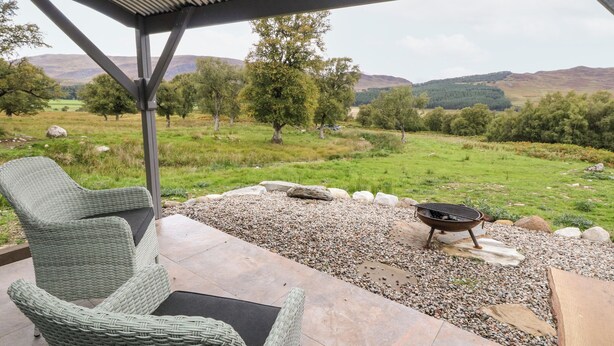 Ein überdachter Außenbereich mit Korbmöbeln und einem Grill, mit Blick auf eine grüne Landschaft mit Bäumen und Hügeln.
