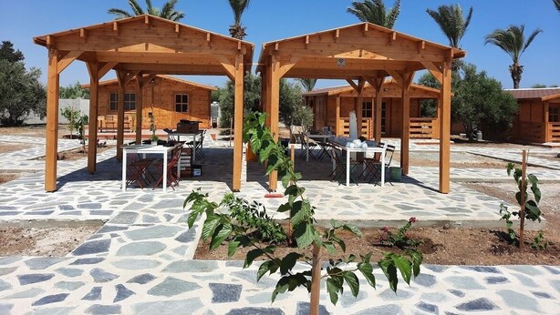 Two wooden pergolas with tables and chairs, set on a stone patio with palm trees in the background.