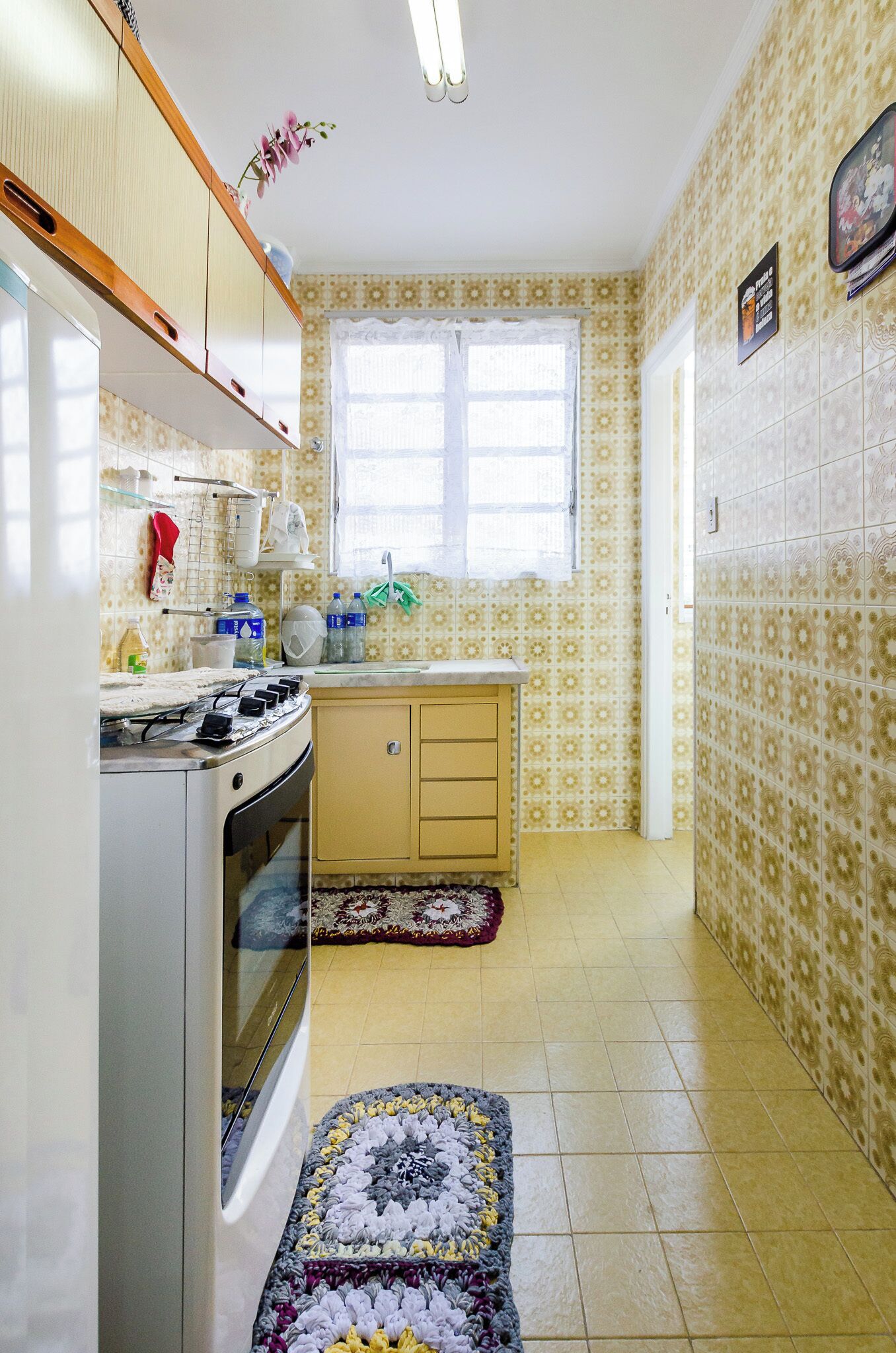 A compact kitchen with a gas stove, oven, and a small countertop. There is a window with a curtain, patterned wallpaper, and a rug on the floor.