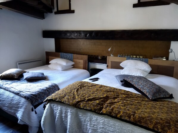 A hotel room with two beds, each with white bedding and multiple pillows. The room has a wooden headboard and a small table with a lamp. - Hostelry (Écully)