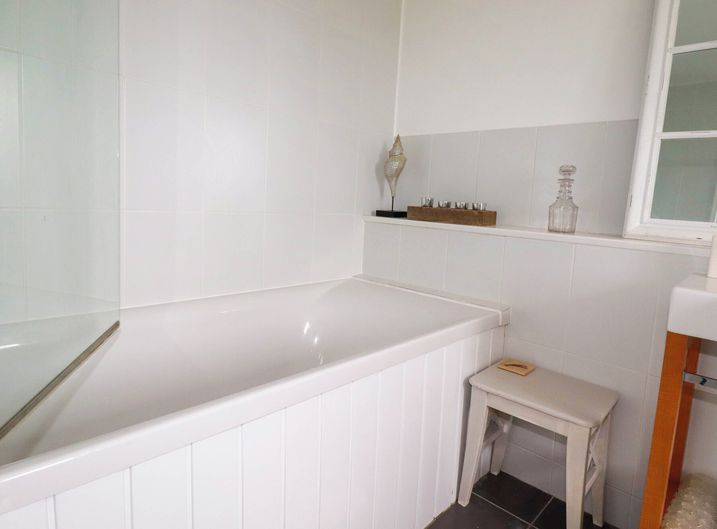A bathroom with a white bathtub, a small white vanity, and a shelf with decorative items.