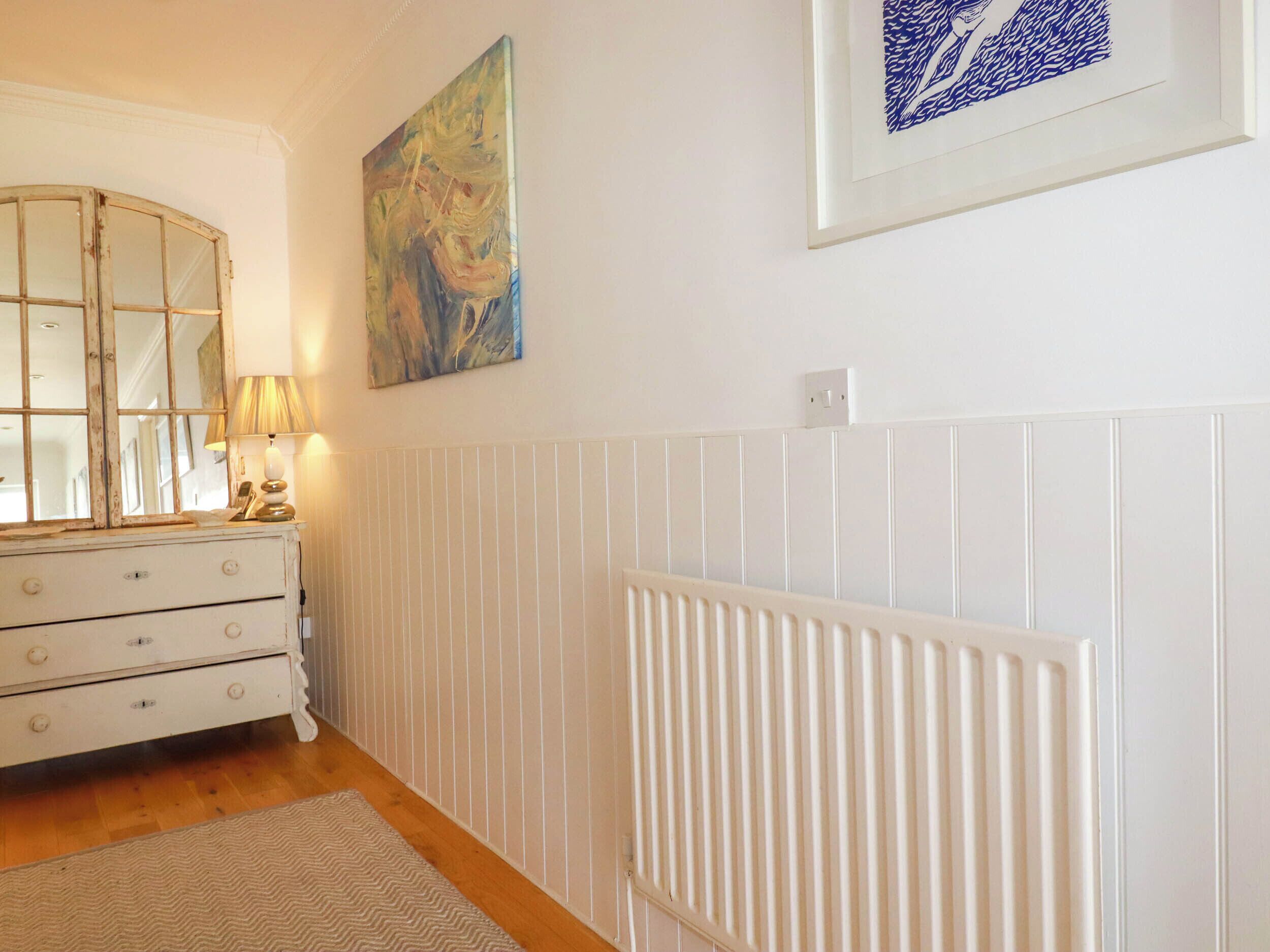 Une pièce avec une commode blanche, une lampe, un radiateur et deux œuvres d’art encadrées accrochées au mur.