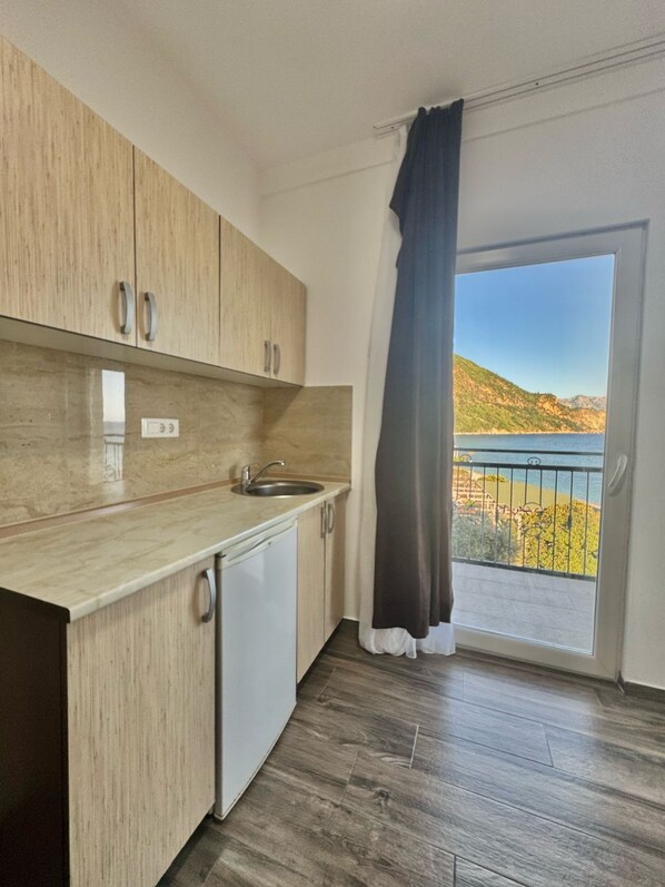 A kitchen with wooden cabinets, a sink, and a dishwasher. There is a large window with a view of a coastal landscape. - Beachfront Hotel Palma Jaz (Prijevor)