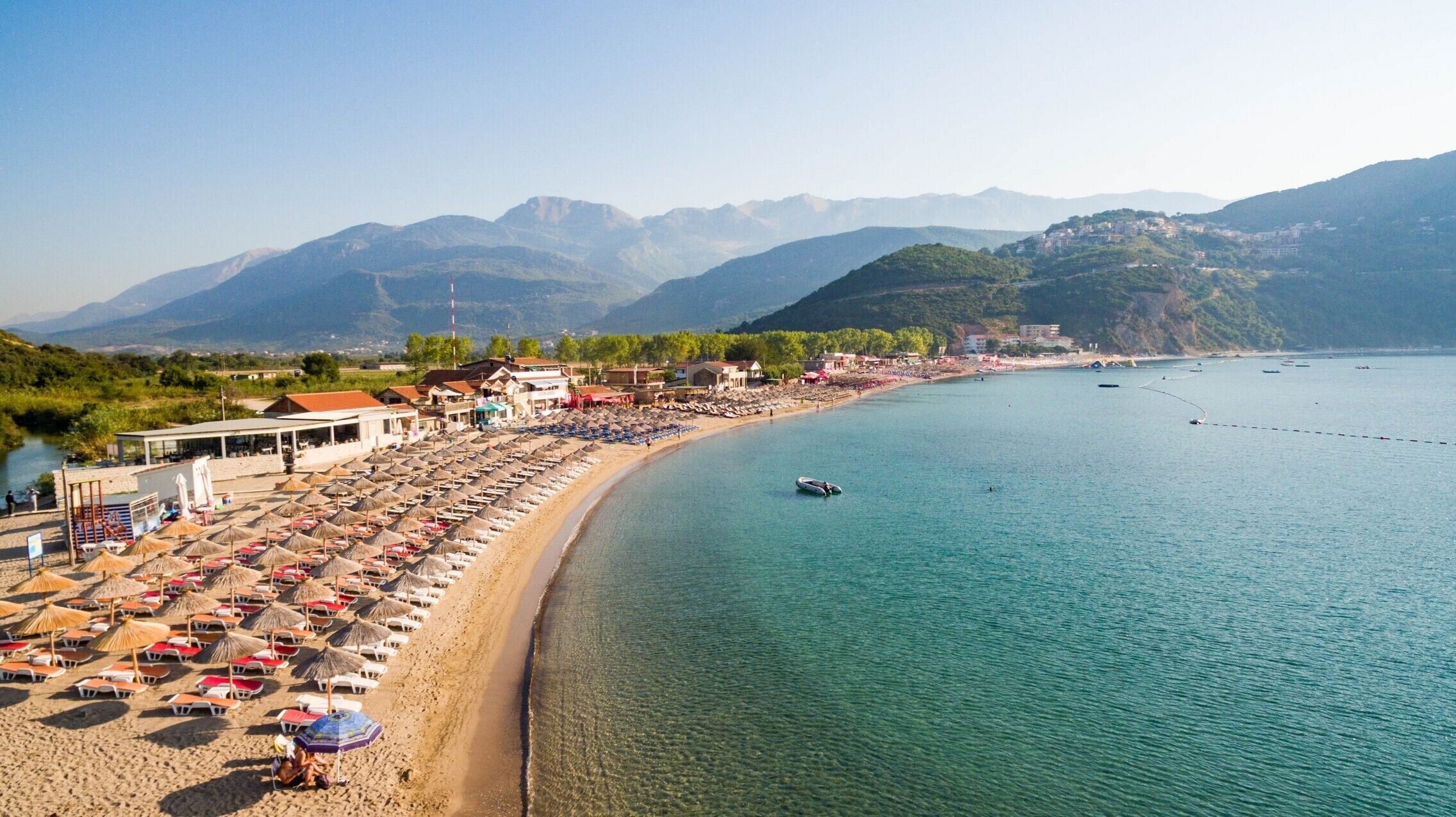 Une plage côtière bordée de rangées de parasols et de chaises de plage, une mer bleue et limpide, et des montagnes en arrière-plan.