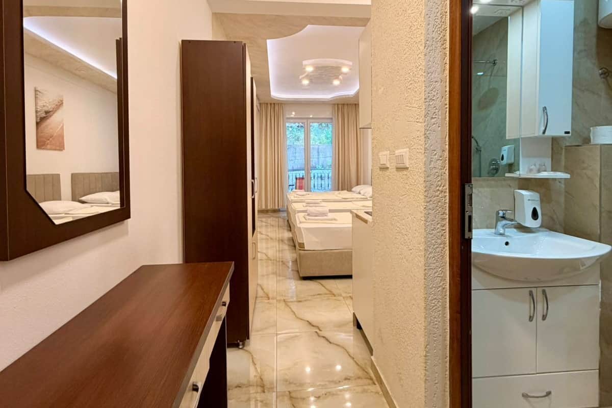 A hotel room with a wooden desk, a large mirror, a bathroom with a sink and cabinets, and a view of the outside.