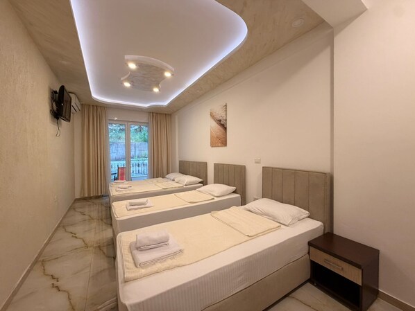 A hotel room with two beds, a small bedside table, a ceiling light fixture, and a view of the outdoors. - Beachfront Hotel Palma Jaz (Prijevor)