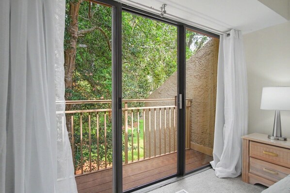 A room with a sliding glass door leading to a balcony with a wooden railing, a small bedside table with a lamp, and a view of greenery outside.
