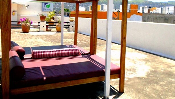 Una terraza en la azotea con una pérgola de madera, zona de estar con cojines, plantas en macetas y vistas de la ciudad.