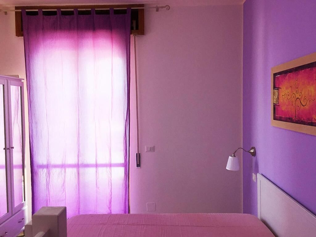 A bedroom with a bed, a window with purple curtains, a wall-mounted lamp, and a painting.
