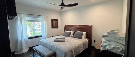 A bedroom with a bed, a ceiling fan, a window with curtains, a bedside table, and a stack of towels.