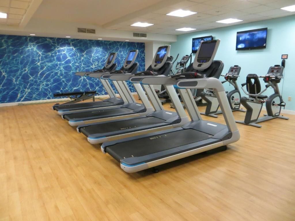 A row of treadmills in a gym with a blue marble wall and a television mounted on the wall.