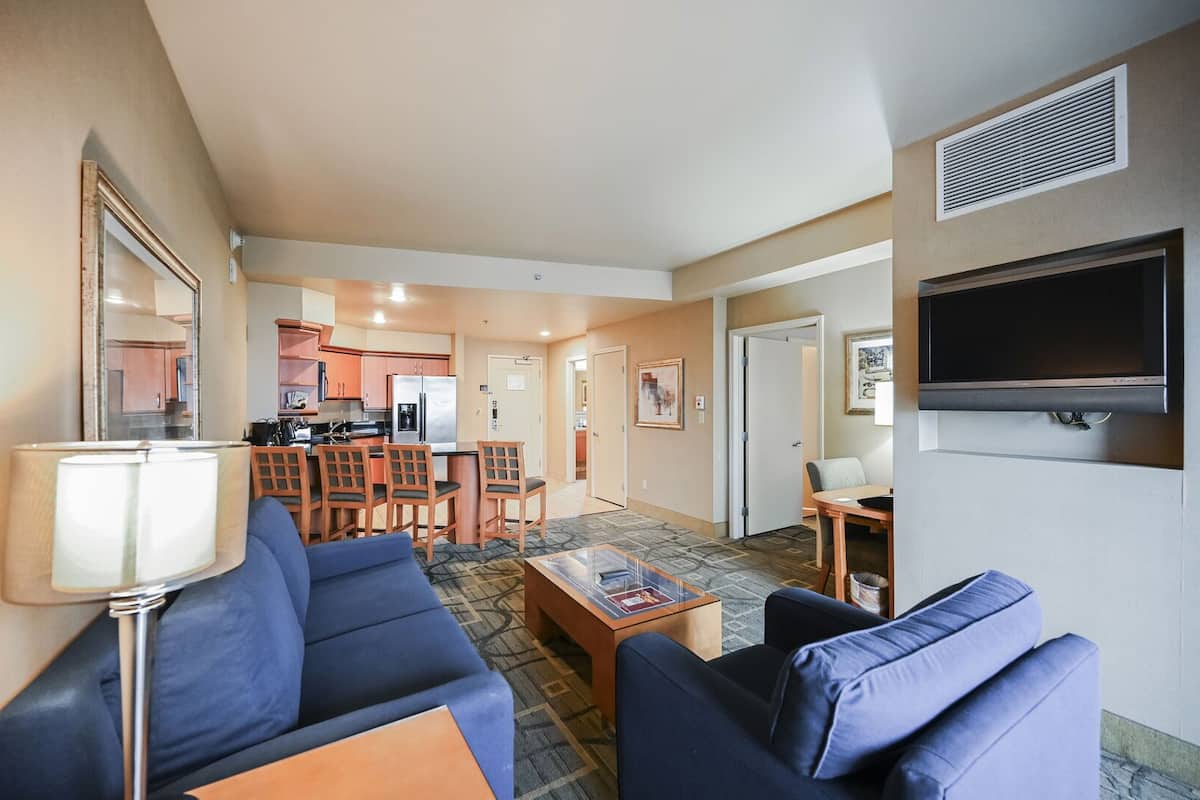 A modern hotel room with a living area featuring blue sofas, a coffee table, and a TV. There is a kitchen area with a refrigerator and a dining table.