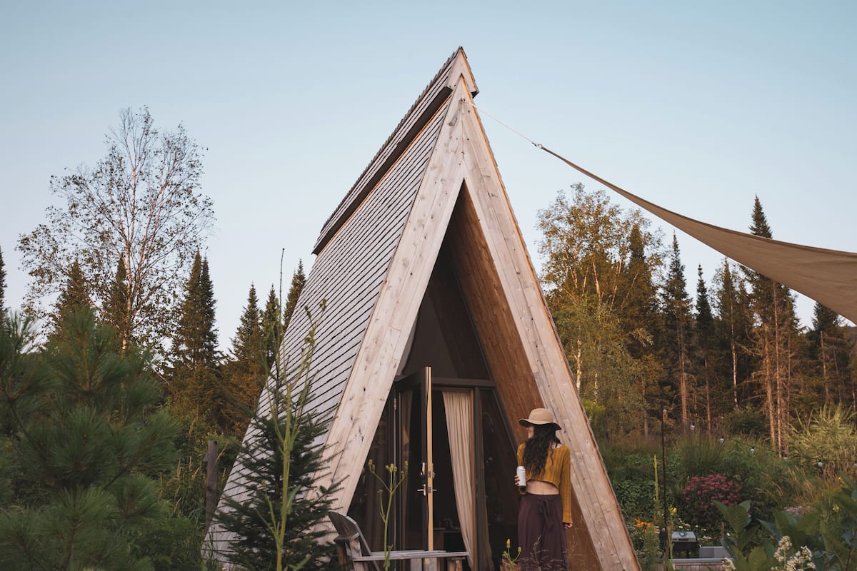 Una cabaña de madera triangular con porche, rodeada de un bosque denso.