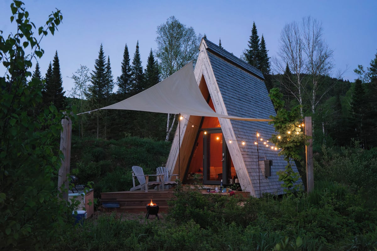 Una cabaña con techo triangular, una terraza con sillas y una guirnalda de luces.