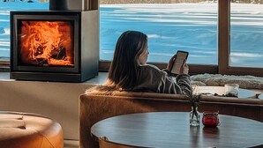 Ett mysigt rum med en öppen spis, ett stort fönster med utsikt över ett snötäckt berg och en person som sitter och läser en bok.