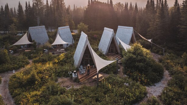 En rad triangulära tält med en träveranda och en sittplats, omgivna av tät skog och kullar i bakgrunden.
