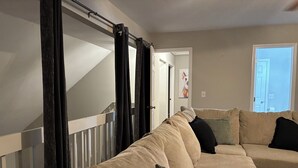 Un saló amb un sofà seccional beix, una escala amb cortines negres i una porta que condueix a una altra habitació.