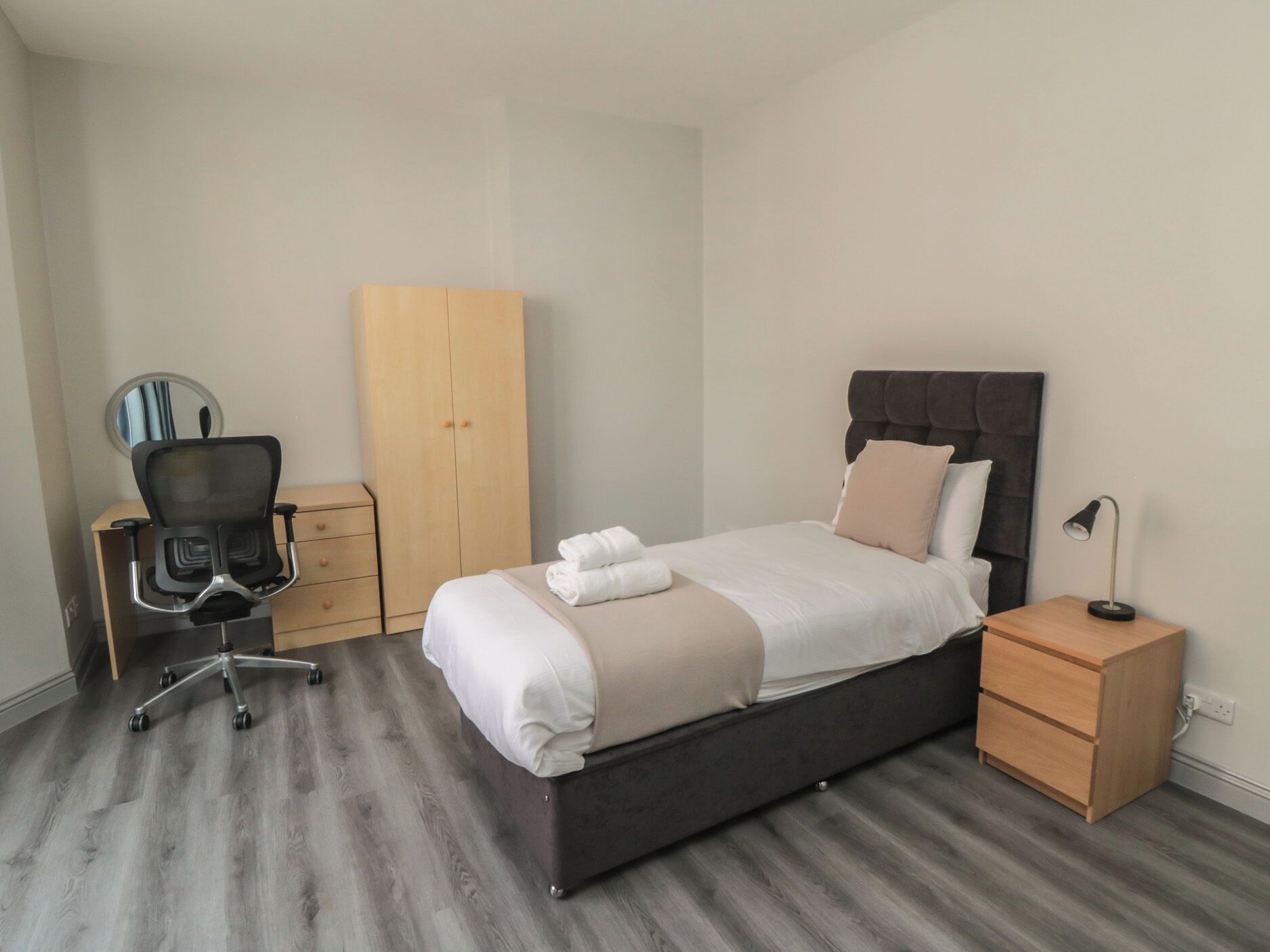 A bedroom with a bed, a desk with a chair, a wardrobe, and a bedside table with a lamp.