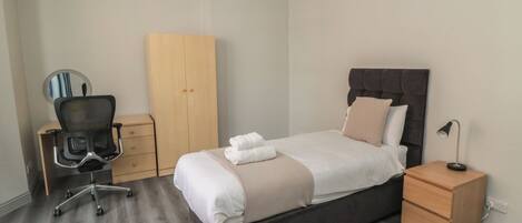 A bedroom with a bed, a desk with a chair, a wardrobe, and a bedside table with a lamp.