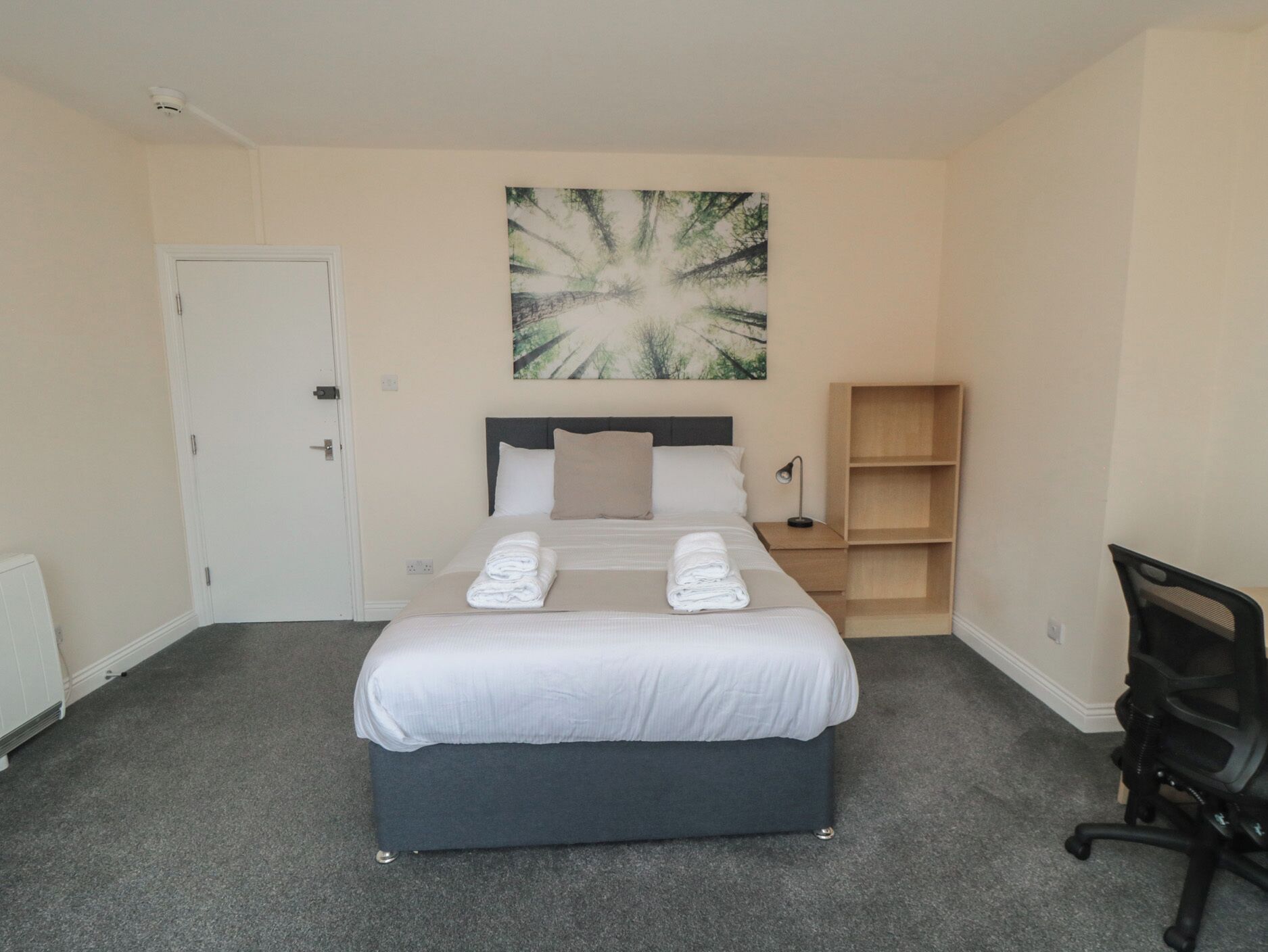A neatly arranged bedroom with a bed, a bedside table, a chair, and a wall-mounted shelf.