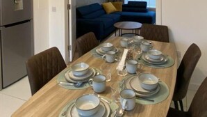 Un coin repas avec une table en bois, dressée pour quatre personnes, entourée de chaises. On y trouve également une cuisine moderne avec un réfrigérateur situé à gauche.