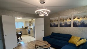 Un salon avec un canapé bleu, un fauteuil jaune et une table basse en bois. On aperçoit un coin repas avec une table et des chaises à travers une porte ouverte.