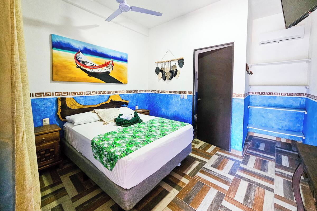 A bedroom with a bed, a ceiling fan, a painting, a door, and patterned flooring.