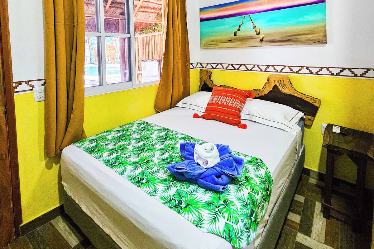 A room with a bed, a ceiling fan, a window with curtains, a painting on the wall, and a wooden headboard.