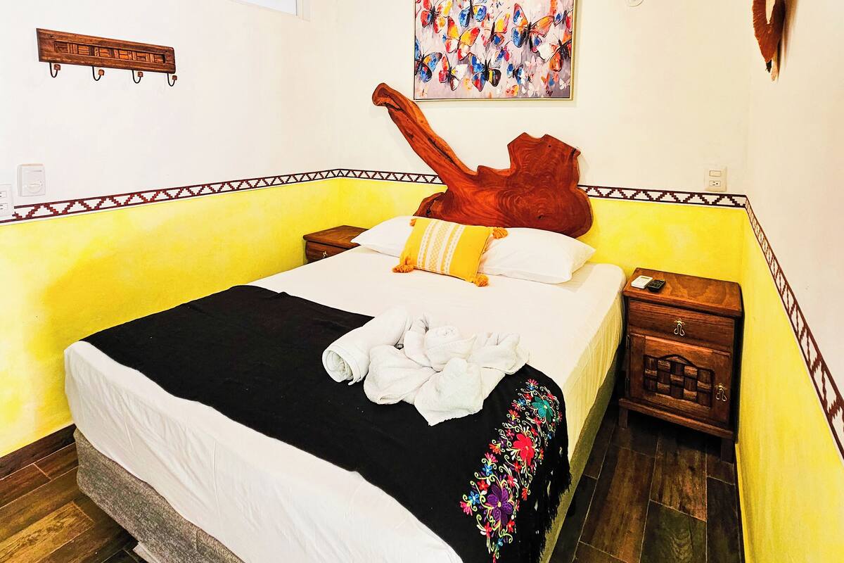 A hotel room with a bed, a ceiling fan, a wall-mounted artwork, a wooden nightstand, and a decorative wall piece.