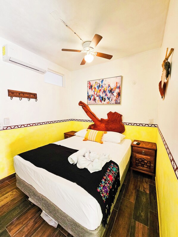 A hotel room with a bed, a ceiling fan, a wall-mounted artwork, a wooden nightstand, and a decorative wall piece. - Hotel Maya Dreams (Bacalar)