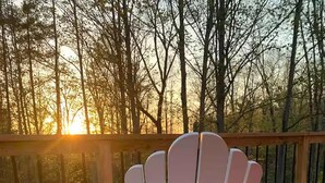 Una terraza de madera con una silla Adirondack blanca y una mesita, rodeada por una baranda de madera y vistas a árboles.