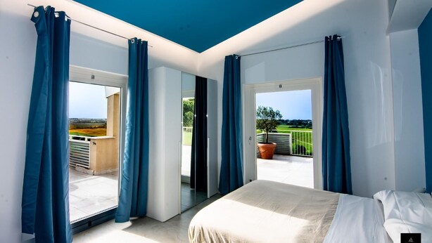 Ein Schlafzimmer mit einem großen Bett, einem Balkon mit blauen Vorhängen und Blick ins Grüne.