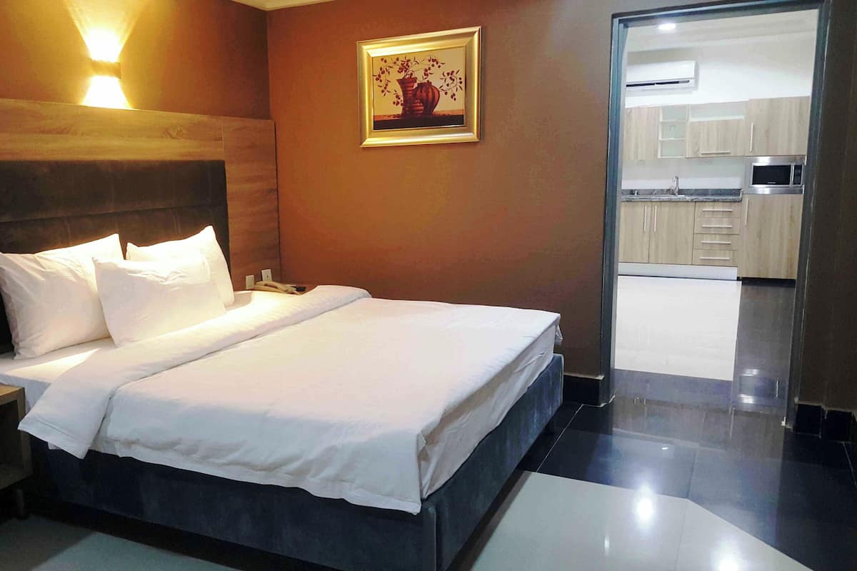A modern hotel room with a large bed, bedside tables, a nightstand, a painting on the wall, and a view of the kitchen.