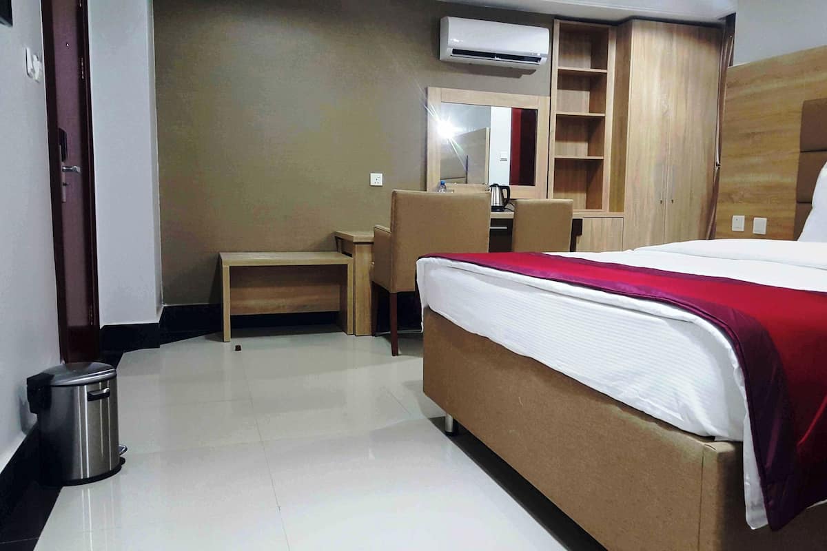 A hotel room with a bed, a desk, a chair, a trash bin, a wardrobe, and an air conditioning unit.