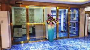 Un área de recepción con suelo de baldosas azules, un mostrador de recepción, un jarrón grande con flores y un cartel que indica el centro de negocios.