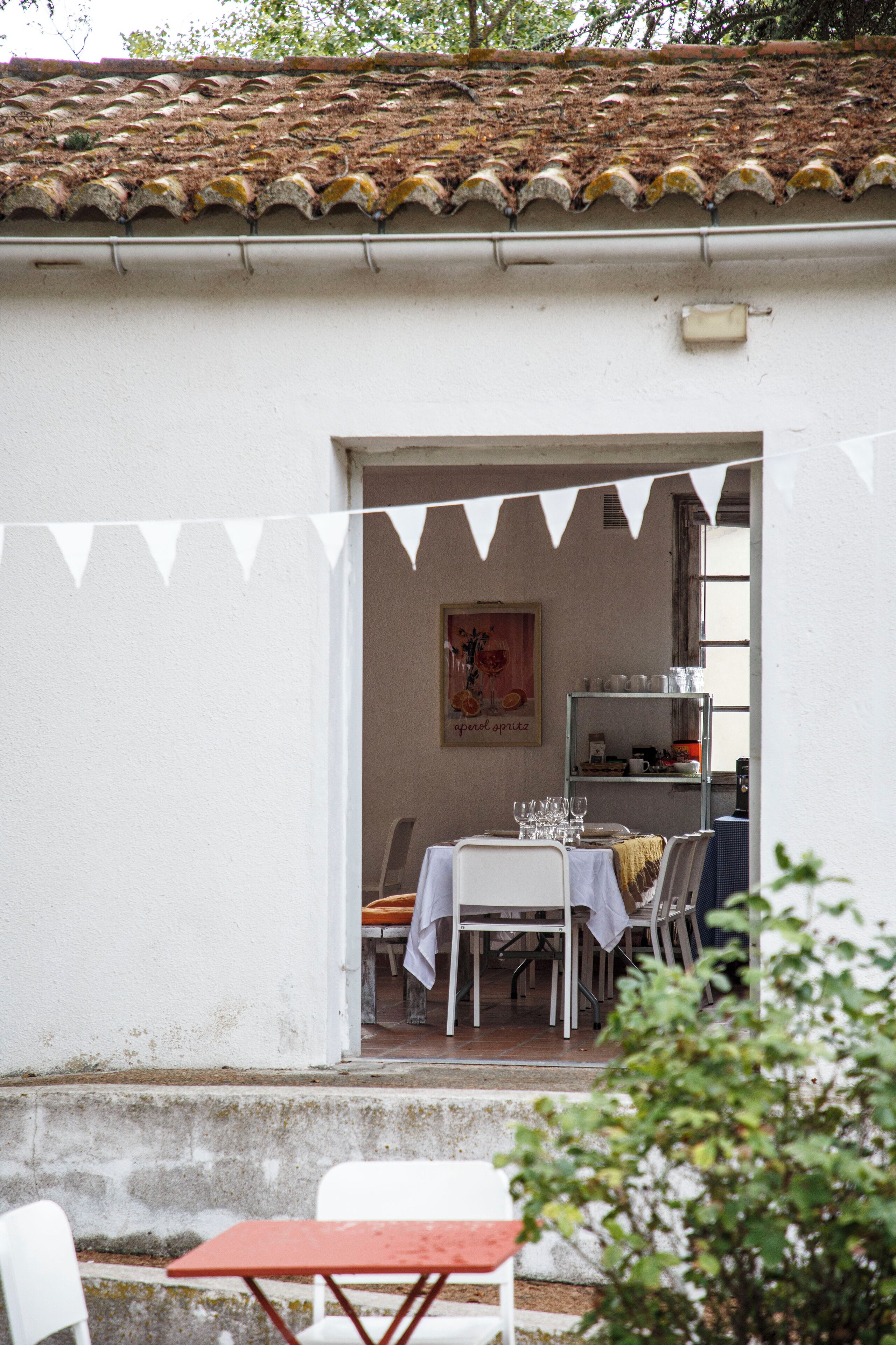 Un espace extérieur aménagé pour les repas, avec une table et des chaises, une étagère où sont rangés de la vaisselle et un toit recouvert de tuiles.