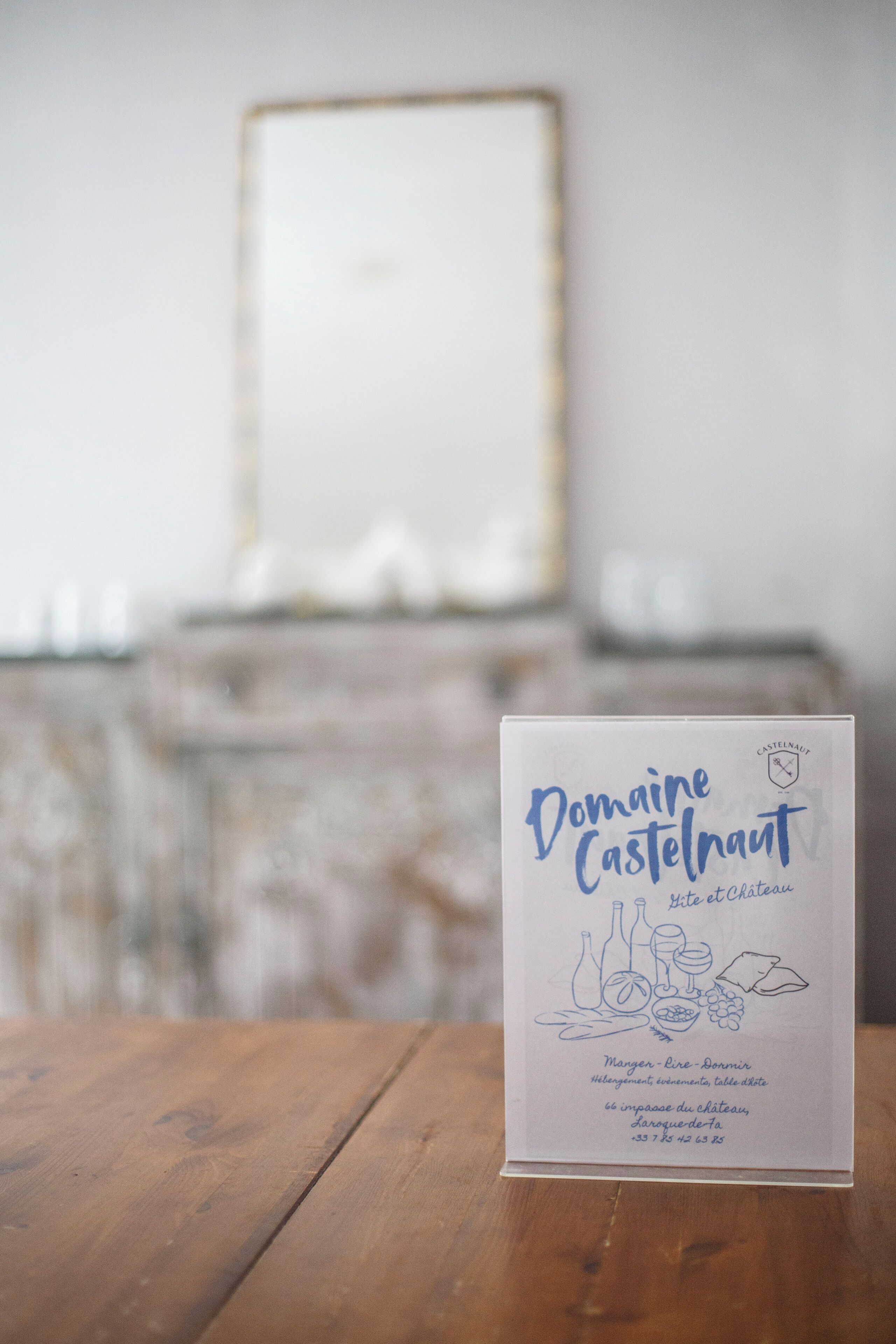 Un menú con el título 'Domaine Castelnaut', con ilustraciones de comida y vino, sobre una mesa de madera en una habitación con fondo desenfocado.