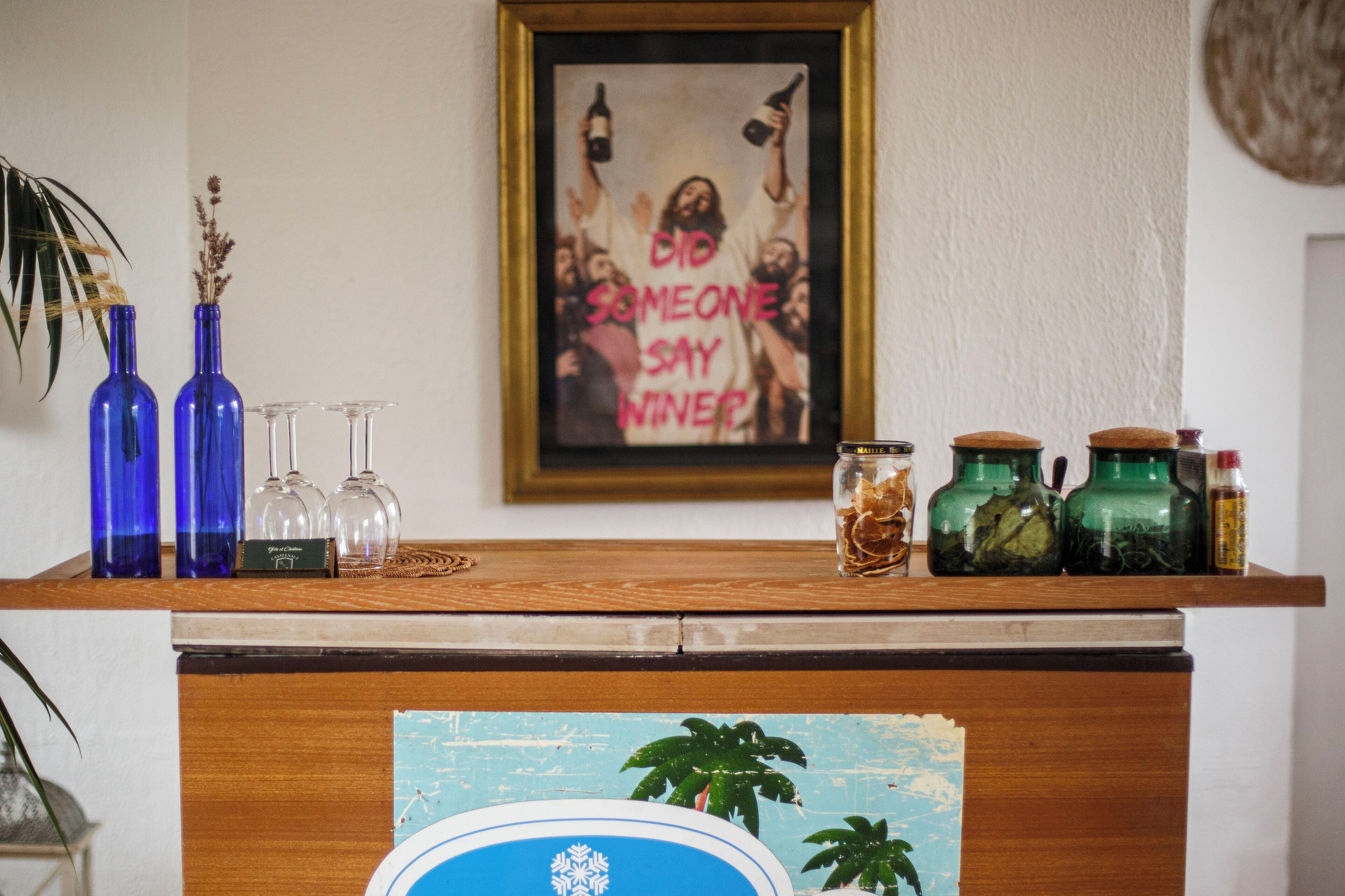 A wooden counter with blue and green glass bottles, clear wine glasses, and a framed poster on the wall.