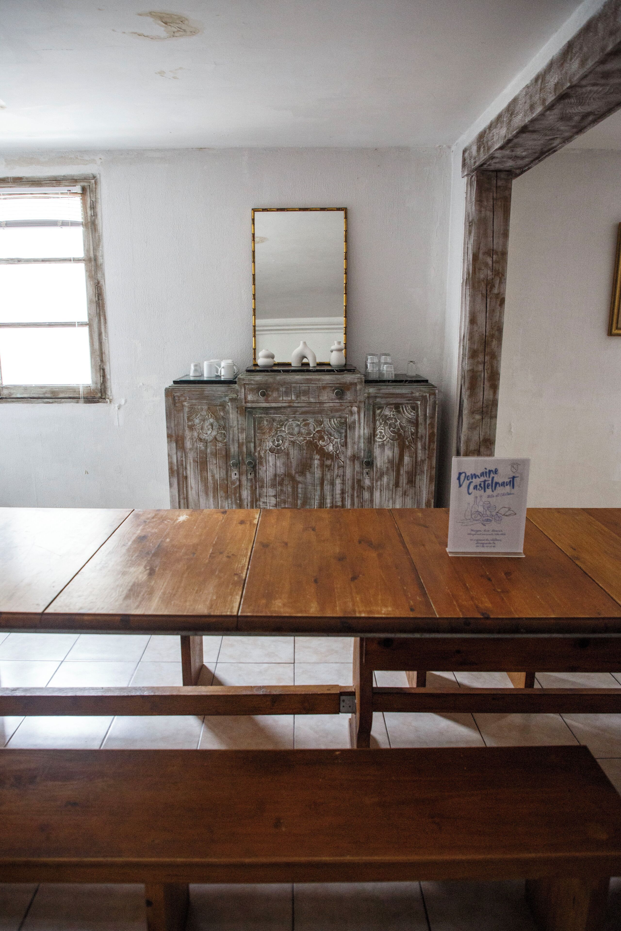 Een houten tafel met banken in een kamer met een rustieke, versleten houten kast en een spiegel aan de muur.