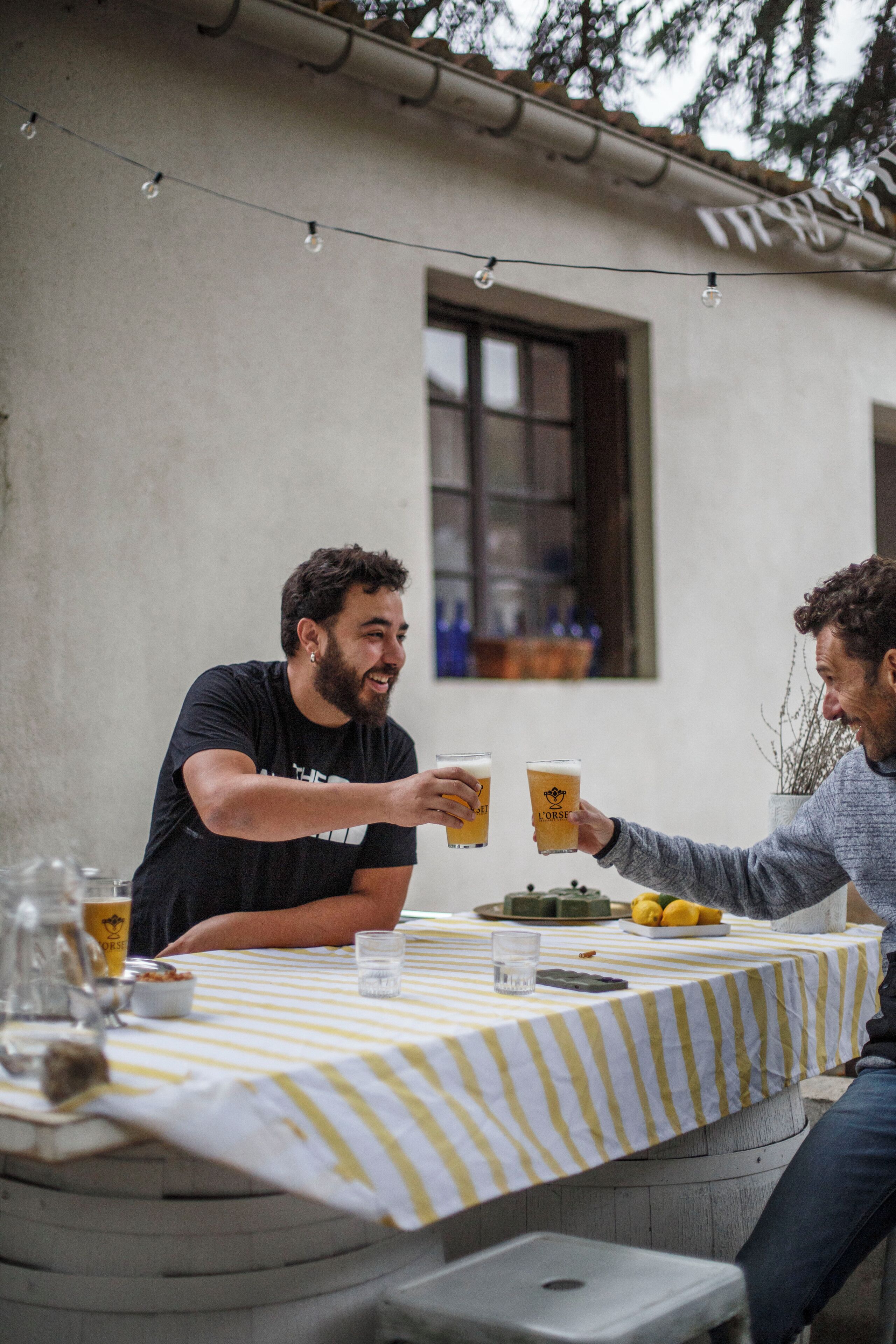 Dues persones brindant a taula amb un llençol rayat, una ampolla i un got de cervesa.
