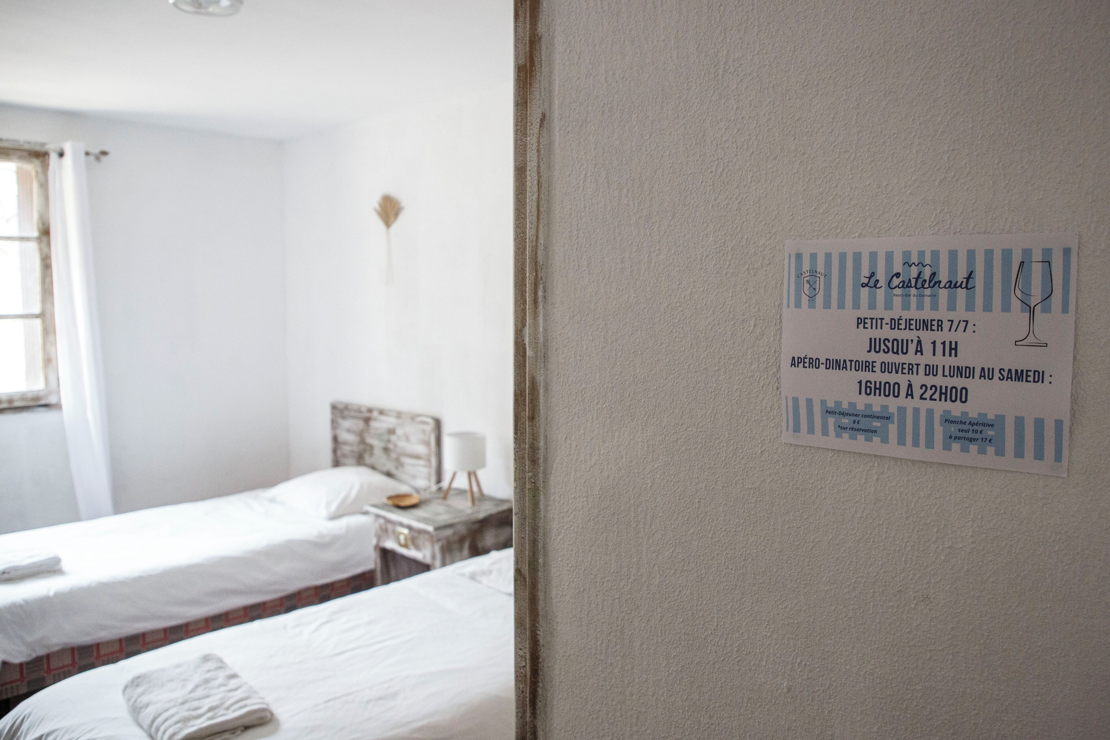 Habitación de hotel con dos camas, una mesita con una lámpara y una pared con un cartel que indica el horario de apertura del restaurante.