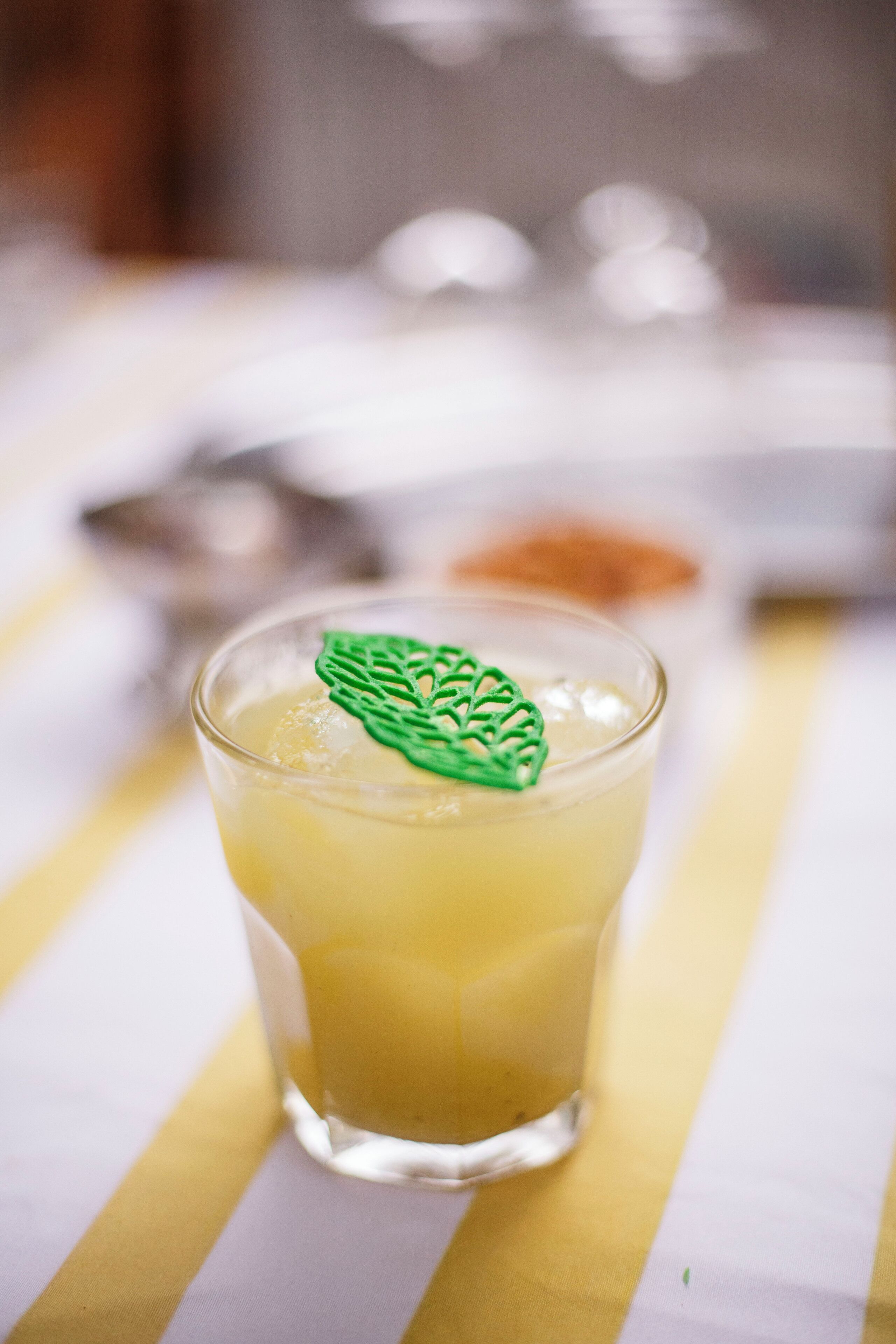 Un vaso de bebida amarilla con una ramita de hoja verde sobre un mantel a rayas.