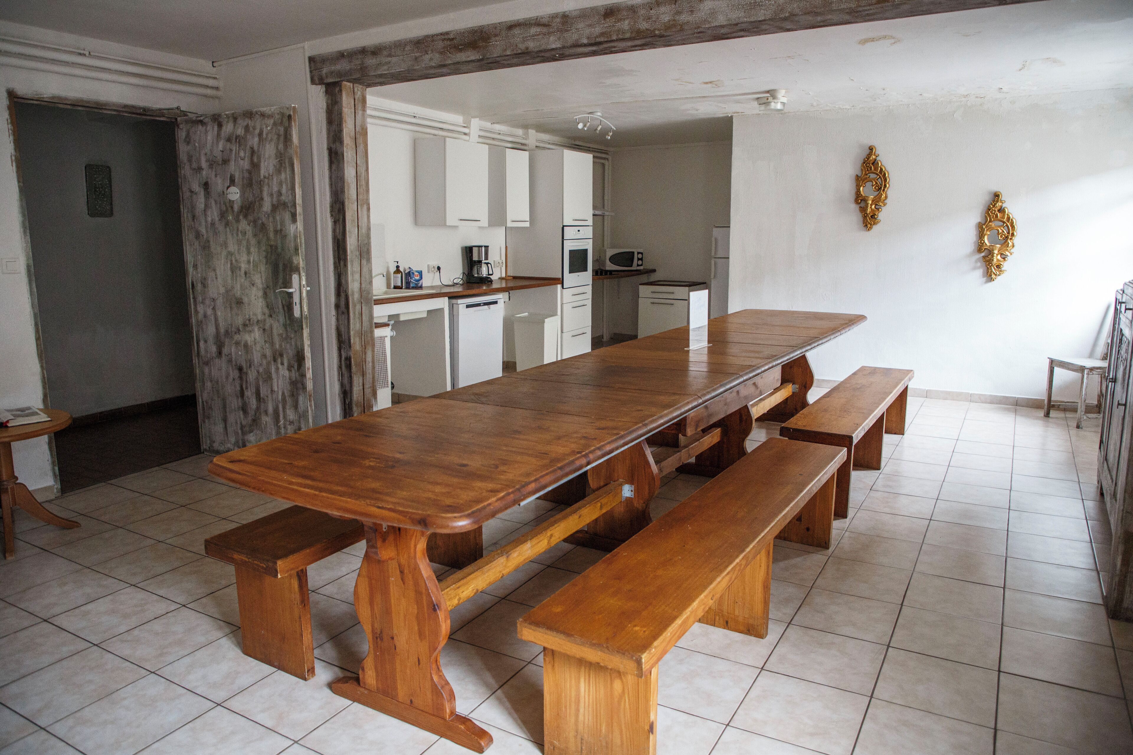 A spacious dining area with a large wooden table and benches, a kitchen in the background, and a tiled floor.