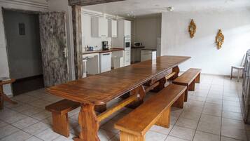 A spacious dining area with a large wooden table and benches, a kitchen in the background, and a tiled floor.