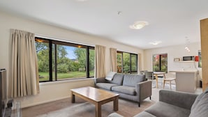Ein modernes Wohnzimmer mit einer Couch, einem Couchtisch und einem Fernseher. Große Fenster mit Vorhängen, eine Küchenzeile und Blick ins Grüne.