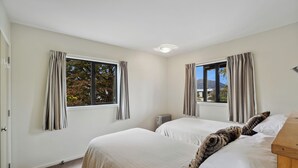 Ein Schlafzimmer mit zwei Betten, großen Fenstern mit Vorhängen, einem Heizkörper und einem Nachttisch aus Holz.